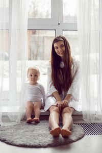 Mother in a white robe sits with a child a blonde daughter at a large window of the house person