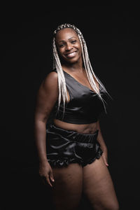 Confident smiling adult african american female with dyed afro braids wearing black silk pajama looking at camera in dark studio