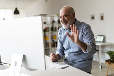 Cheerful senior businessman waving hand on video call through desktop computer in home office