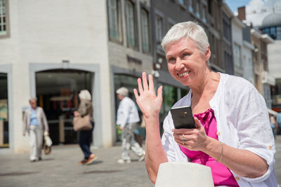 Sixty year old caucasian woman in stylish summer clothes waves to a passerby, holds smartphone