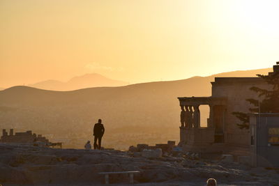 Rear view of man standing by historic building by mountains against clear sky during sunset