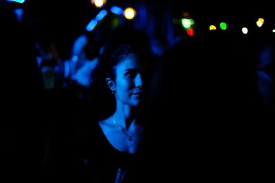 Young woman looking away while standing against illuminated lights at night