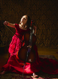 A small beautiful child in long red dress plays cello in window light