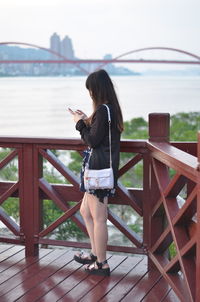 Side view of woman using phone while standing on footbridge in city