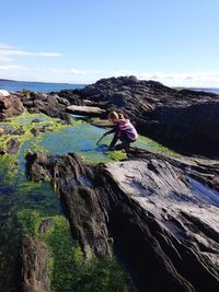 Side view of woman crouching by moss covered tide pool against sky