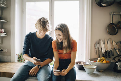 Teenage boy showing mobile phone to female friend while sitting at kitchen counter