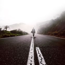 Man walking on road in front of mountains against sky during foggy weather