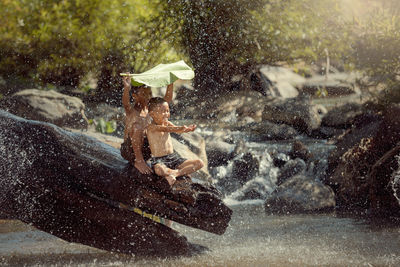 Happy shirtless brothers sitting on log by lake during rainfall