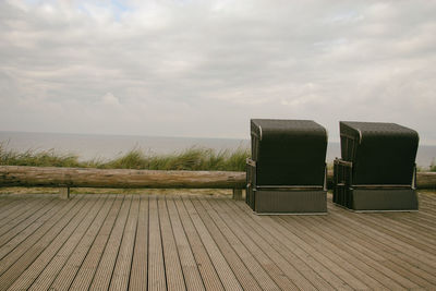 Hooded beach chairs on wooden boardwalk by sea against cloudy sky
