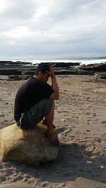 Man sitting on rock at beach against sky