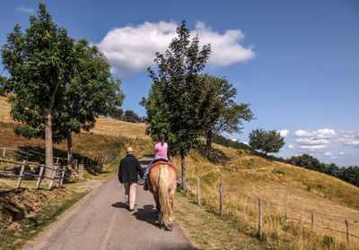 Girl riding horse with man on road amidst trees against sky