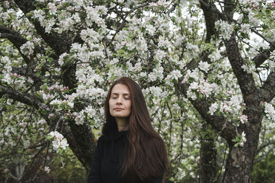 Portrait of beautiful young woman standing against white flowering apple tree