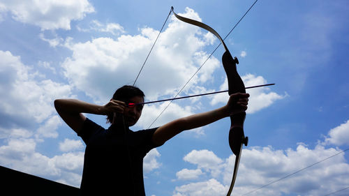 Low angle view of woman holding bow and arrow while standing against sky
