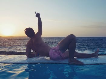 Rear view of shirtless man lying at poolside against sky during sunset