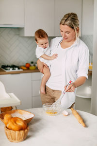 A young mother holds her baby in her arms, whips the eggs with a whisk and prepares the dough.