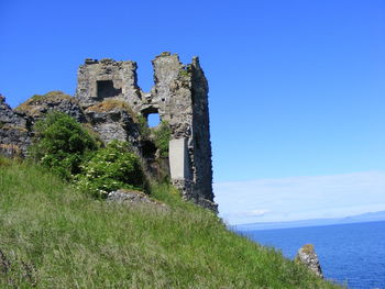 Old abandoned built structure on grassy field by sea against sky during sunny day