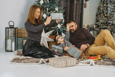 Family with child playing near christmas tree. child unpacking gifts, middle aged parents enjoy