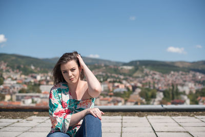 Portrait of young woman with hand in hair sitting against sky