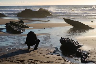 Rear view of man crouching at beach during sunset