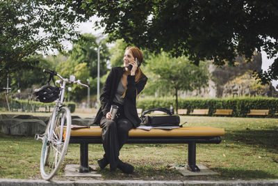 Happy businesswoman talking on mobile phone while sitting by bicycle at park
