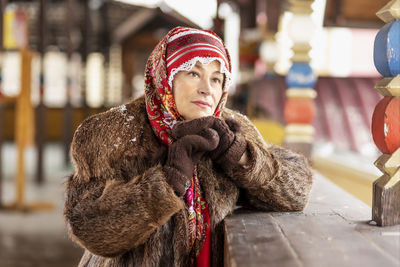 A slavic woman in a national colored scarf,a fur coat and  mittens on the porch of a wooden house