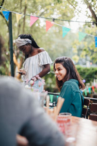 Smiling young woman looking away while sitting on chair at table in balcony during dinner party