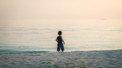 Rear view of baby boy standing on shore at beach against sky during sunset
