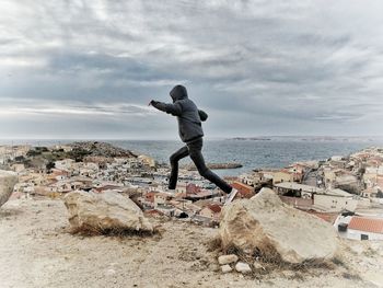 Man jumping over rock at beach against cloudy sky