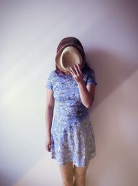 Woman covering face with hat against white wall