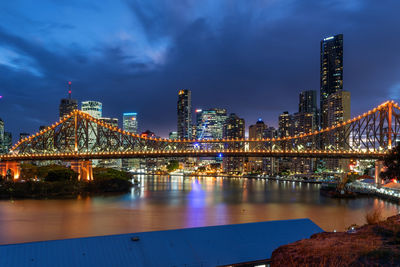 Illuminated bridge over river by buildings against sky at night