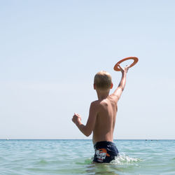 Rear view of shirtless boy standing in sea against clear sky