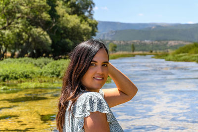 Portrait of attractive young woman with long hair, standing outdoor in front of river on a sunny day