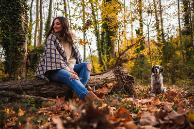 Wide shot of a woman sitting on a log with her dog in a forest.