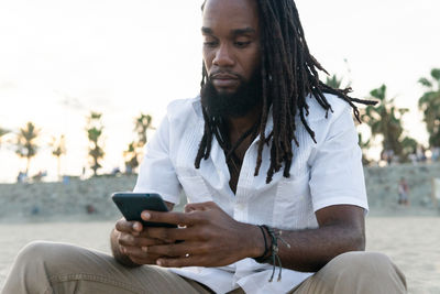 Concentrated african american male with dreadlocks browsing mobile phone while sitting on sandy beach against blurred background of trees