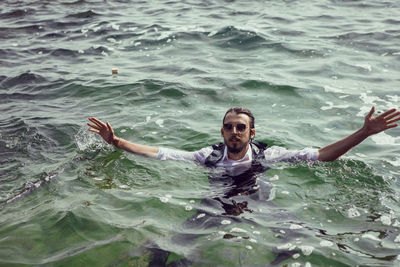 Man with a beard and sunglasses in clothes a vest and a white shirt swims in the sea among the rock