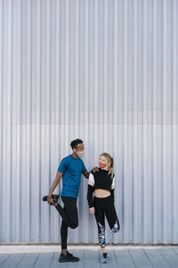 Male and female sportsperson with protective face mask doing warm up exercise while standing against wall during pandemic