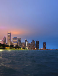 Illuminated buildings by sea against sky at dusk