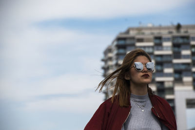 Confident young woman standing against cloudy sky in city