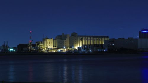 Illuminated buildings by river against blue sky at night