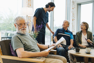 Portrait of retired senior man sitting with newspaper on armchair by friends and caretaker at elderly nursing home