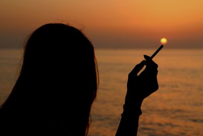 Silhouette woman holding cigarette by sea during sunset