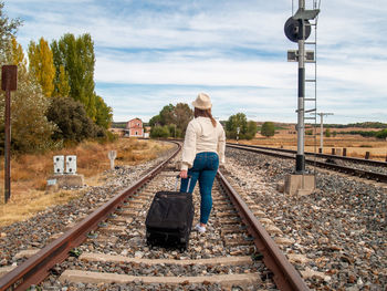 Rear view of woman walking with luggage on railroad track against sky