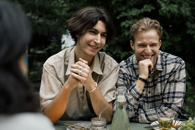 Smiling young man with hand on chin sitting by male friend during dinner party