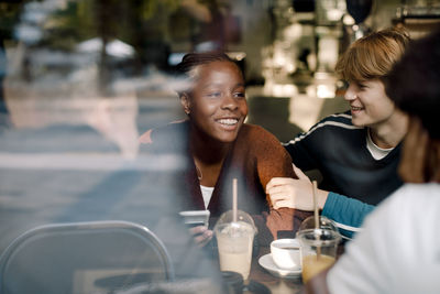 Smiling teenage girl with friends at cafe seen through glass window