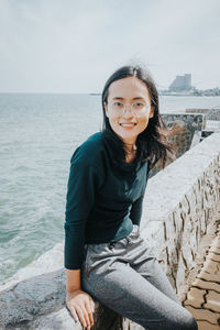 Portrait of smiling young woman sitting by sea on retaining wall