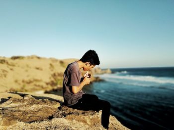 Man sitting on rock by sea against clear sky