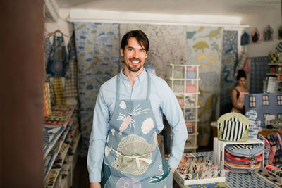 Portrait of smiling male owner standing with hand on hip while colleague working in fabric shop