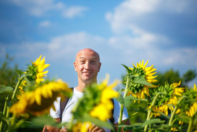 A cheerful bald man in a field of blooming yellow sunflowers against a blue sky stands smiling