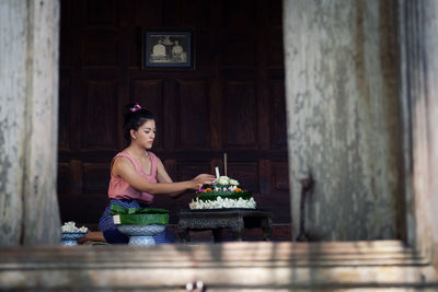 Young woman arranging flowers on table at home seen through window