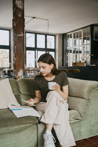 Female using smart phone and having coffee while sitting with legs crossed on sofa in studio